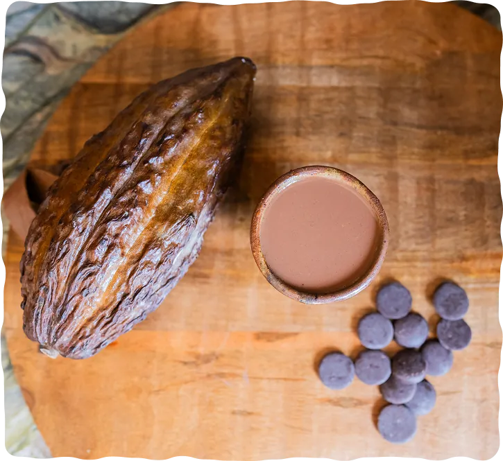 Whole cacao pod beside a cup of prepared ceremonial cacao and cacao discs on a wooden board